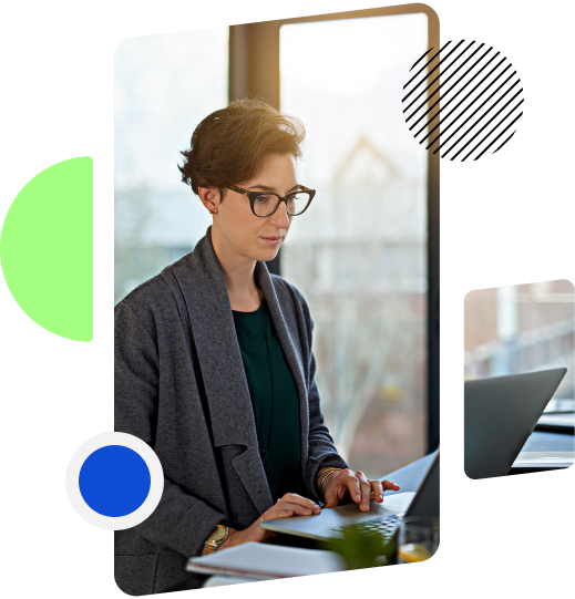 A woman wearing glasses is standing at a desk, focused on working on a laptop in a bright office setting.